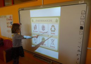 Dziewczynka stoi pod tablicą interaktywną i wskaźnikiem pokazuje z pośród innych znak recyklingu.